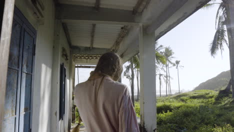 Tall-Russian-brunette-opens-entrance-door-to-a-dark-abandoned-house-and-walks-around-the-corner-to-a-complete-view-of-a-windy-beach-filled-with-palm-trees-and-soaks-in-the-sun