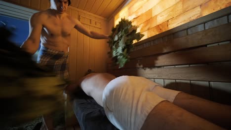 man lying on wooden bench in spa with masseur doing massage with  oak broom. client in finn sauna indoors with professional.