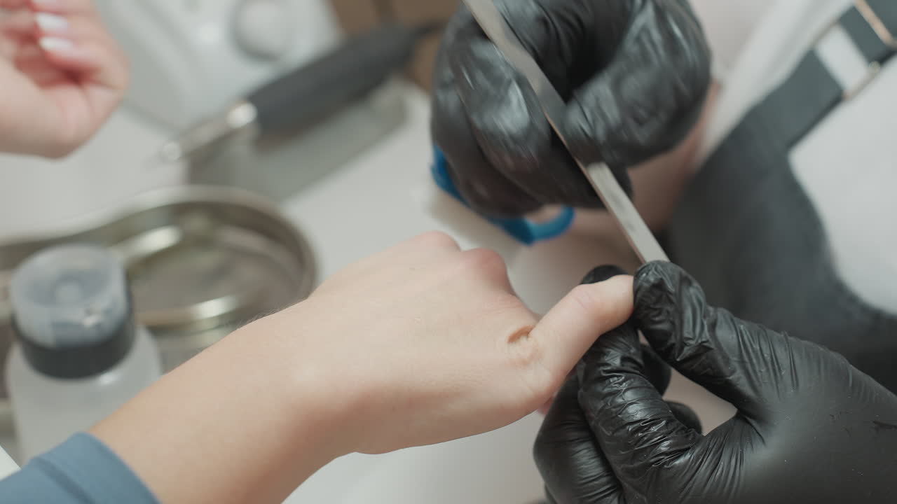 Close-up view of nail technician wearing black gloves filing client nail and using blue brush to clean nail particles, with manicure tools, containers, and equipment visible on white table surface
