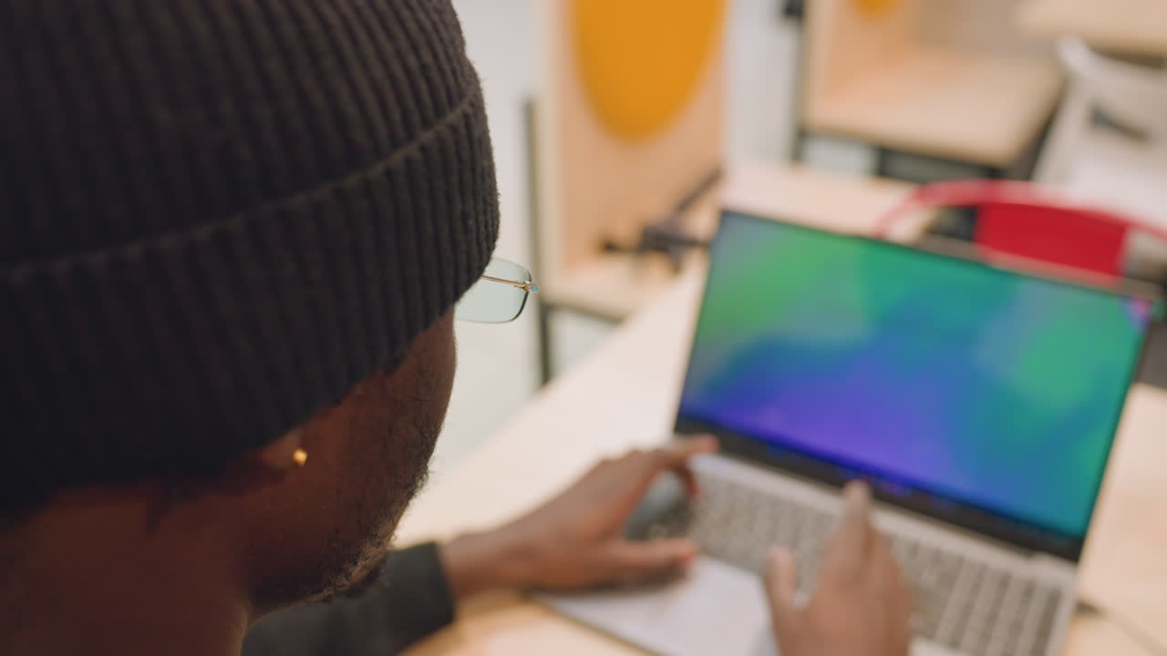 Sort udvikler ved bærbar computer i studie. Nærbilleder over skulderen viser sort mand iført hue og ørering, fokuseret på farverig gradient-skærm, mens han taster og navigerer på trackpad.