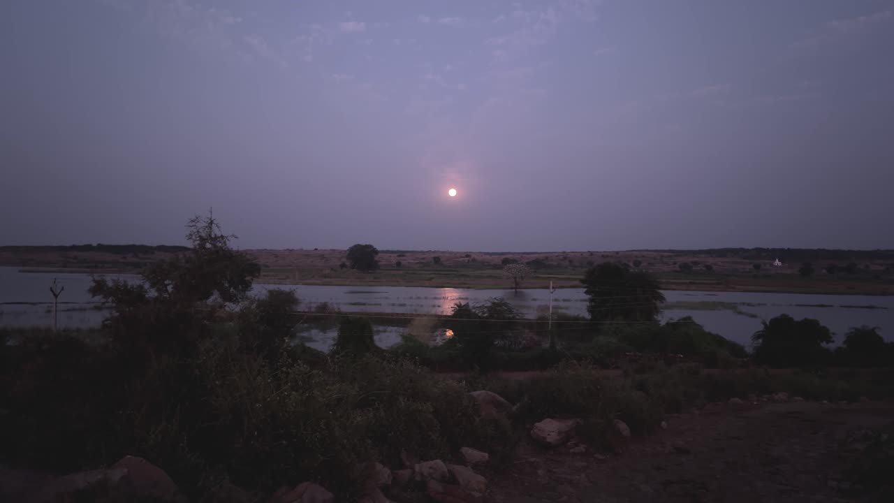 Full moon rise over a lake with water reflection during dawn time in rural india