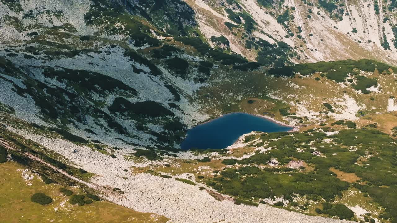 Blue glacial lake in the sunlight close up shot, nested between green pastures and mountain rocks, aerial tracking shot