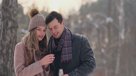 beautiful and stylish couple man and woman hipsters in a coat and scarf drink tea from a thermos in the winter forest after a walk. love story