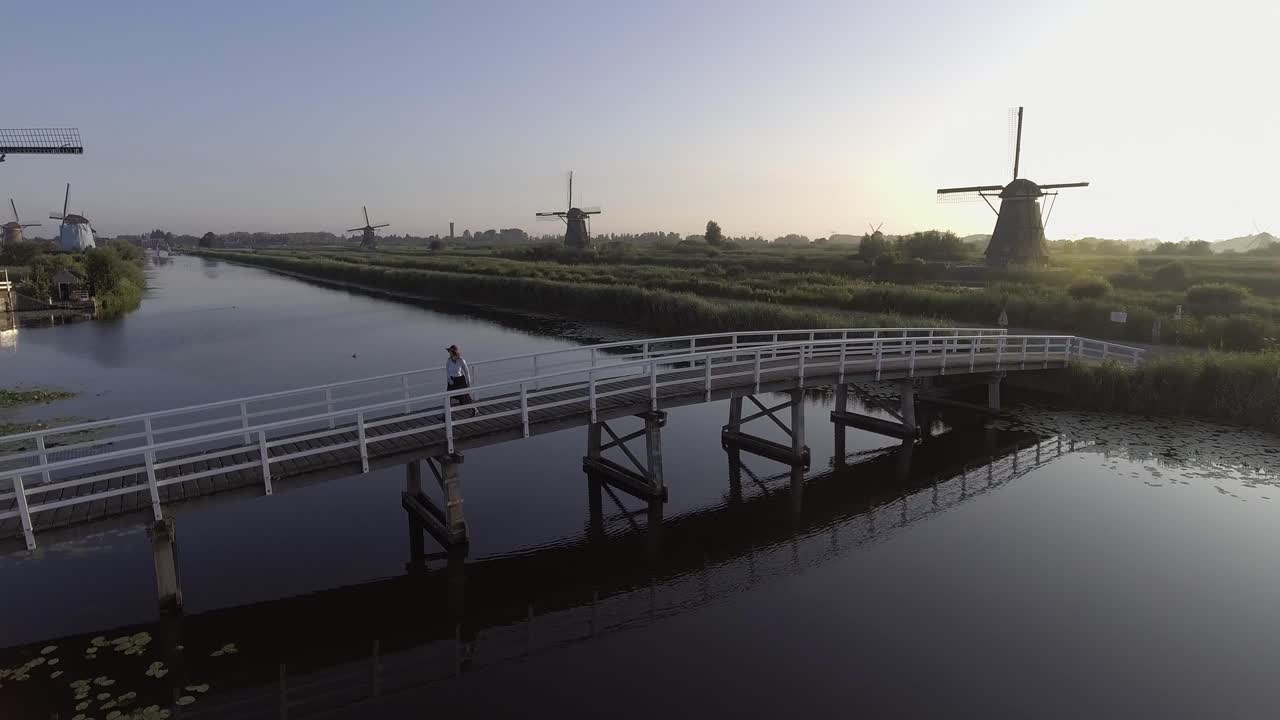 일출 동안 네덜란드의 네덜란드 풍차를 바라보며 다리 위를 걷고 돌아선 소녀 주변을 오른쪽으로 패닝하는 드론 샷