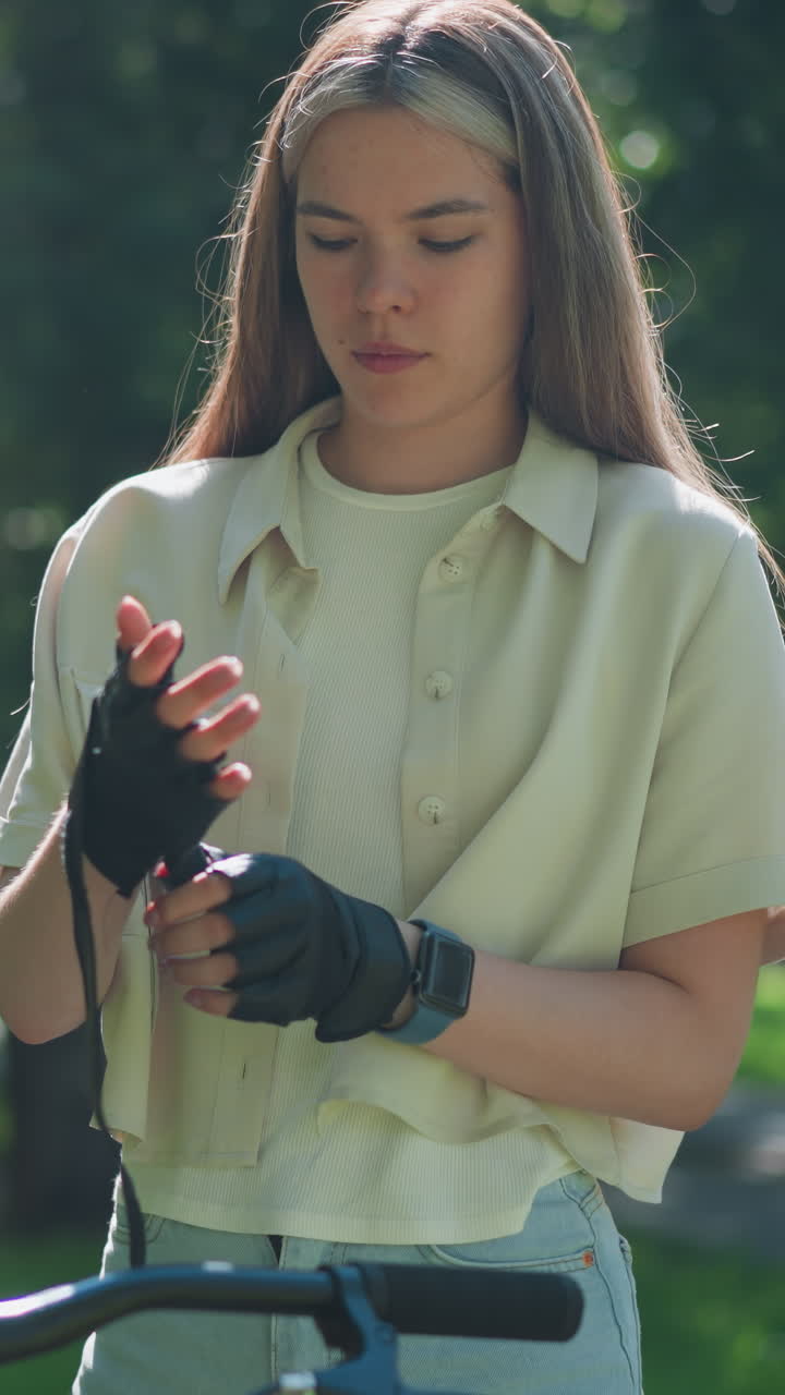 Young woman stands beside her bicycle, carefully adjusting biker glove on her right hand, focused expression on her face, sunlight filters through trees