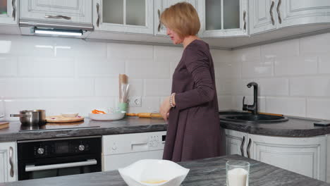 mujer en la cocina moderna preparándose para hornear, apareciendo contemplativa mientras recoge un twister, un cuenco blanco con huevos rotos y un vaso de azúcar sentado en la encimera