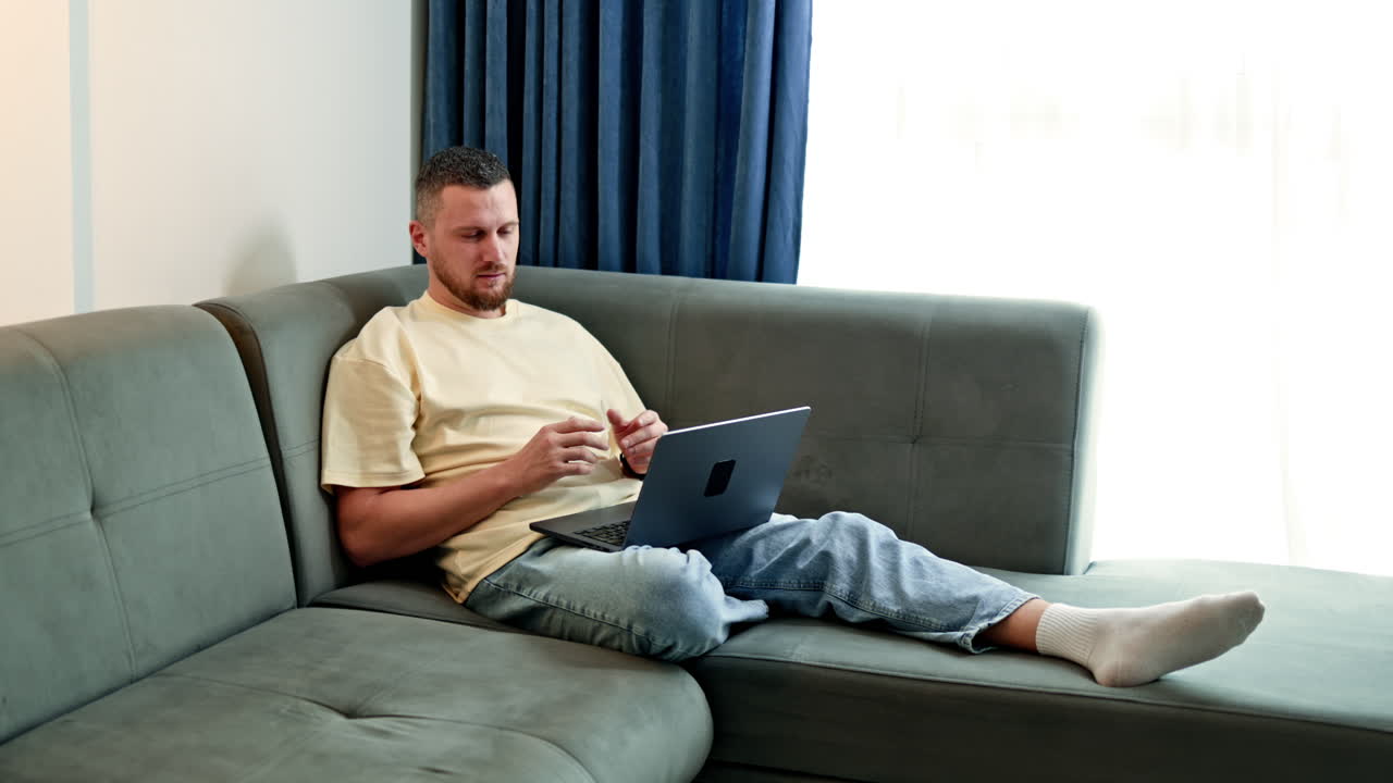 Man sits on sofa working on laptop in living room during afternoon light with window open and curtains drawn