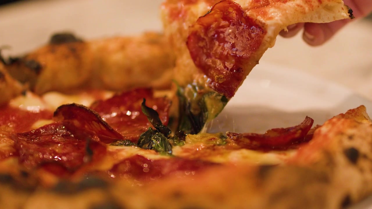 Primo piano di una mano che solleva una fetta di pizza al salame piccante con formaggio fuso e basilico.