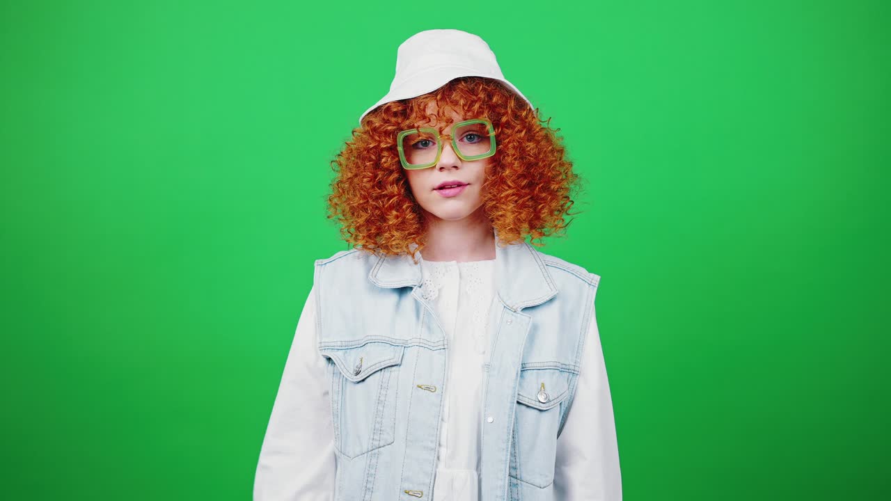 안경과 파나마 모자를 입은 젊은 은 머리 곱슬머리 여성은 패배자 손 제스처, 녹색 크로마 키 배경을 보여줍니다.