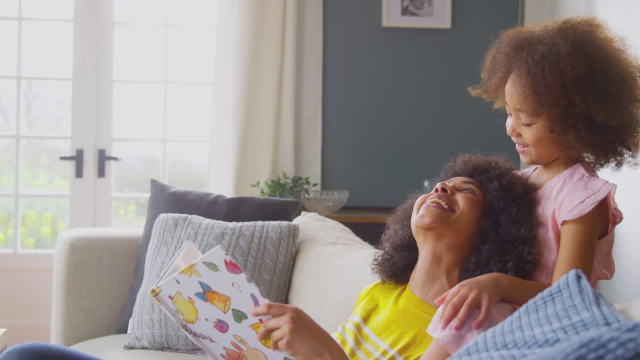 Mother And Daughter Relaxing On Sofa At Home Reading Book Together