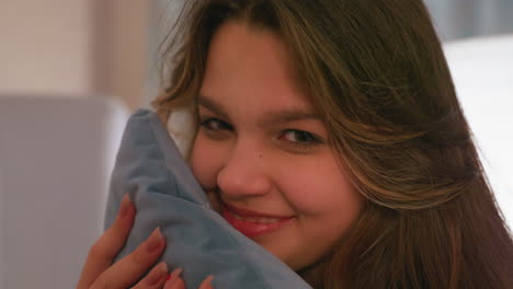 young woman with long brown hair smiles gently while holding soft blue pillow close to her cheek, cozy indoor atmosphere with warm lighting