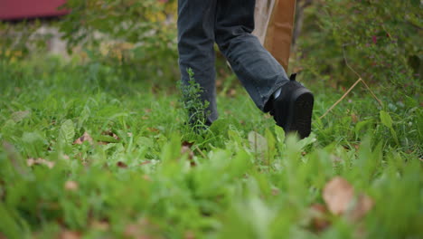 leg view of boy in dark jeans and black sneakers walking through vibrant greenery, holding brown paper bag, surrounded by lush plants and foliage in outdoor natural setting