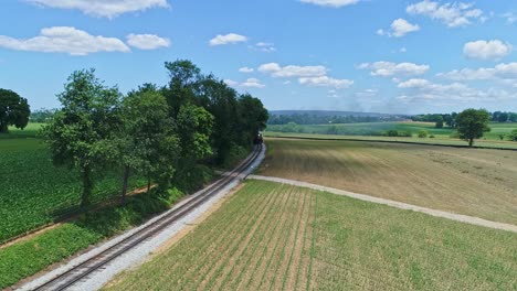 an aerial view of a steam engine puffing smoke and steam with passenger coaches traveling on a single track turn a curve thru trees and farmland countryside on a beautiful cloudless spring day