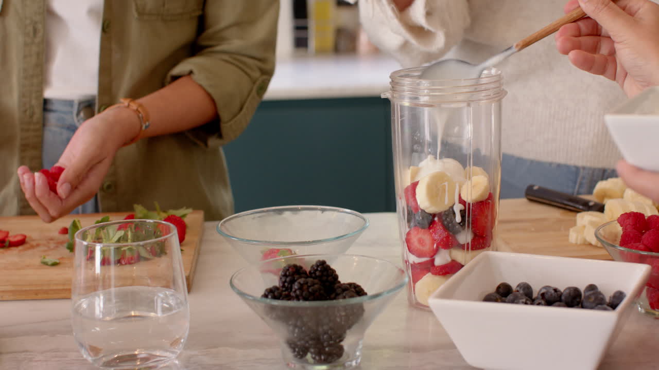 Preparing smoothie, female friends adding fresh fruits in blender in kitchen