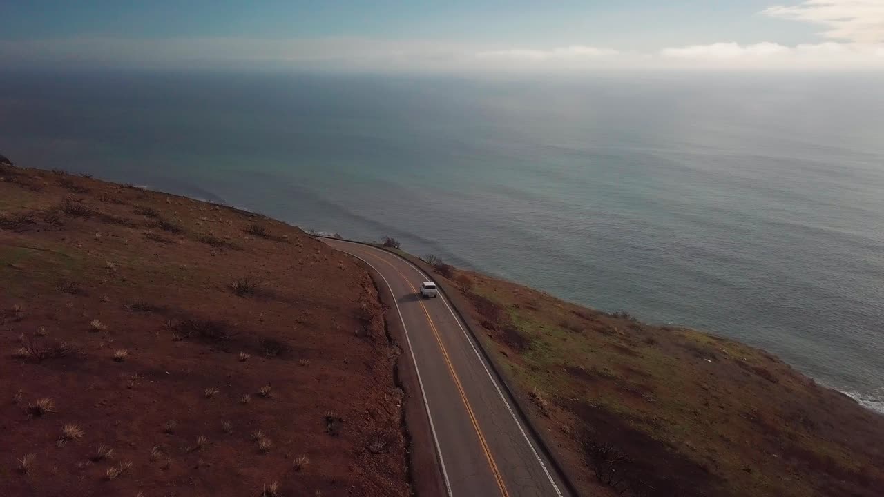 Aerial shot following white SUV along windy mountain road with ocean in the background. 4K