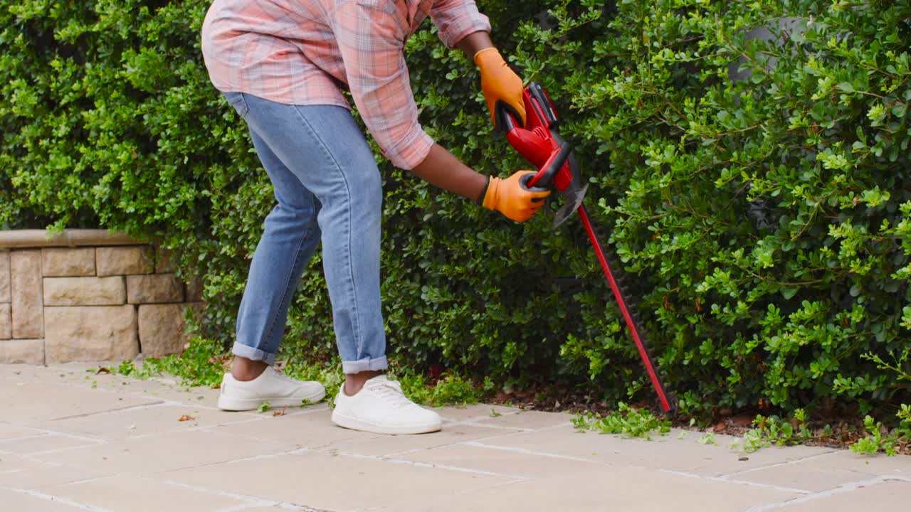 African American man walking along paved walkway using red trimmer in orange gloves removing growth