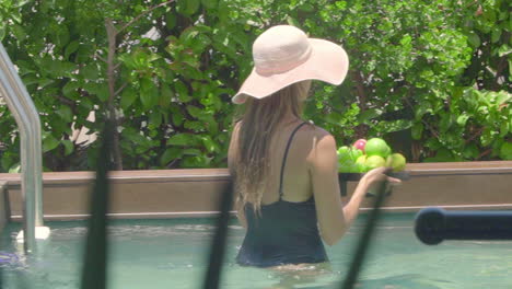 a woman in a black bathing suit and a straw hat carrying a basket of fruit over her private swimming pool