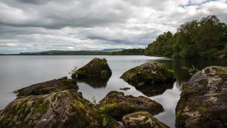 Lapso-De-Tiempo-Del-Lago-Con-Grandes-Rocas-En-Primer-Plano-Y-Bosque-En-La-Distancia-En-Un-Día-Nublado-De-Verano-En-El-Paisaje-Rural-De-Irlanda