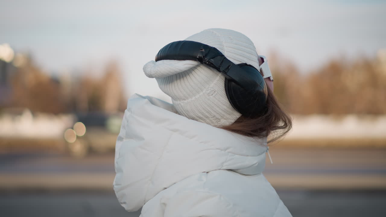 Close up view of dancer smiling with joy, dressed in white winter coat, beanie, large headphones, and sunglasses while dancing in park under soft sunlight, with faint cars passing in background
