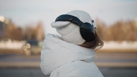 close up view of dancer smiling with joy, dressed in white winter coat, beanie, large headphones, and sunglasses while dancing in park under soft sunlight, with faint cars passing in background