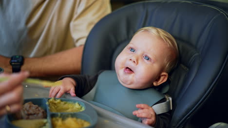blond baby with a blue bib is fed from spoon. caring mom giving purred food to her dear son.
