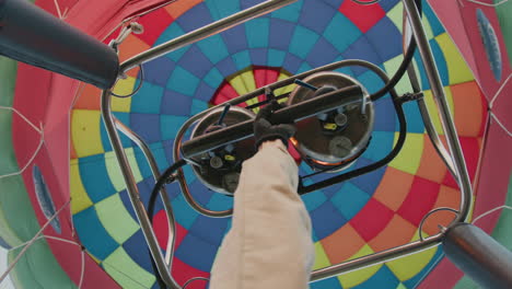 close up of gloved hand reaching up to grip burner controls inside metal frame under bright colorful hot air balloon canopy showing circular patchwork pattern of panels with blue sky background