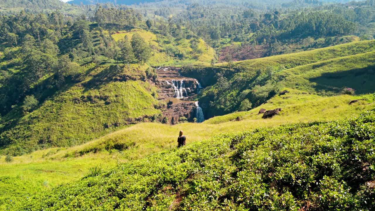 Plongez dans le charme paisible de Saint Clairs, Nuwara Eliya, Sri Lanka, alors qu'une femme marche avec grâce à travers les plantations de thé d'un vert éclatant, avec les emblématiques cascades de Saint Clairs qui dévalent
