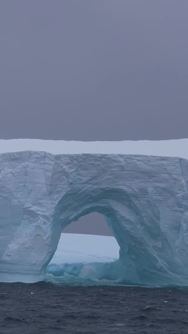 Vertikale Ansicht eines Eisbergs und von Meereswellen, die an einem kalten, nebligen Tag auf Eis brechen.
