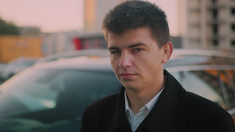 man standing close to car with stern look, blurred background shows parked cars and distant building at dusk, portraying confident business professional