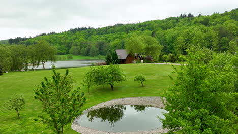 a serene park landscape with a small pond, trees, and a cozy cabin in the background, surrounded by green hills