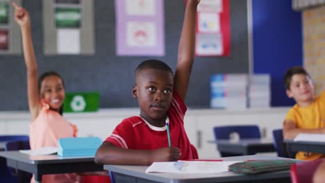 Diverse-schoolchildren-sitting-in-classroom-raising-hands-to-answer-questions-during-lesson