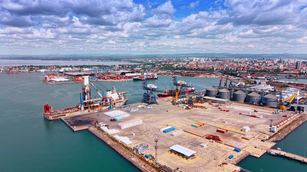 Puerto de Burgas, Barco Perforador, Terminal de Contenedores, Silos, Ciudad. Un barco perforador atraca en la concurrida terminal portuaria de Burgas junto a grúas de contenedores, silos y barcos.