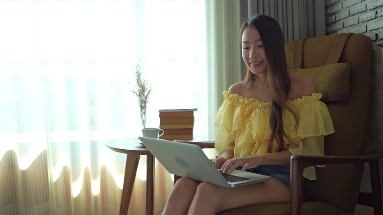 A young attractive Asian woman sitting in comfortable chair types on her laptop