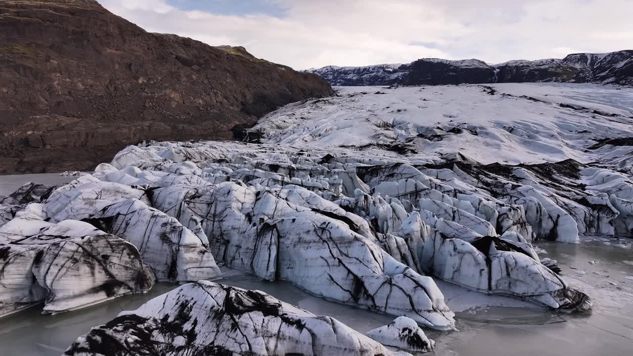 Aerial view of the Sólheimajökull glacier tongue, part of the Mýrdalsjökull glacier in Iceland, showcasing striking ice formations streaked with volcanic ash and surrounded by rugged mountains.