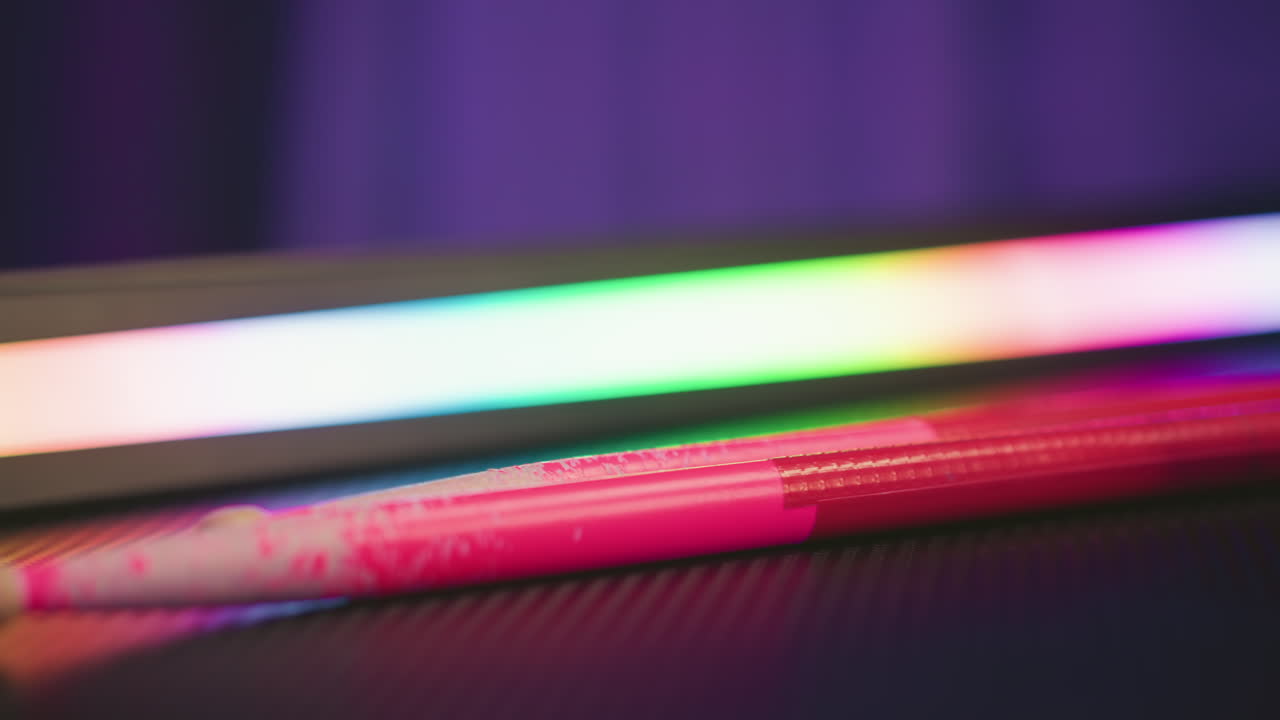 Close up pink drum stick resting on dark textured surface near glowing multicolor LED light bar with blurred vibrant reflections highlighting contrast between musical gear and ambient studio lighting