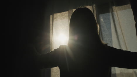 Silhouette-of-a-woman-who-opens-blackout-curtains.-It-is-illuminated-by-the-bright-sun-from-the-window.-Back-view