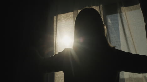 silhouette of a woman who opens blackout curtains. it is illuminated by the bright sun from the window. back view