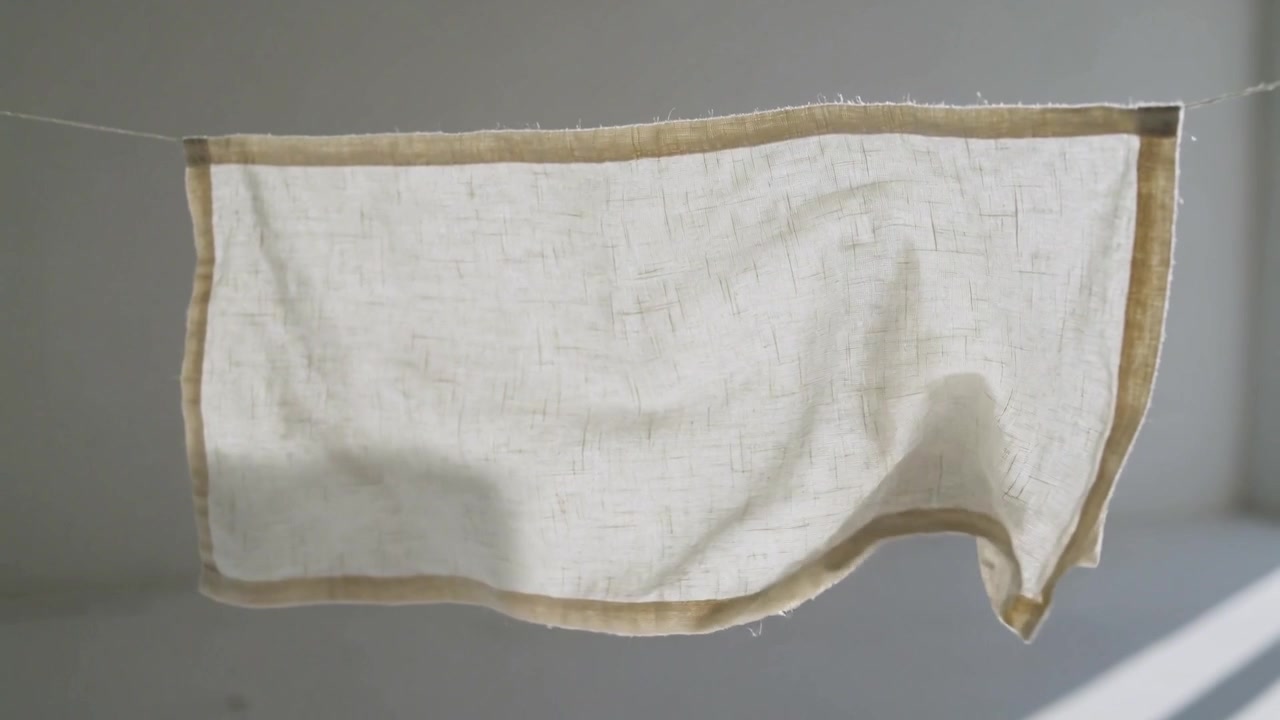 Unrolling linen towel from gentle breeze on clothesline in sunlit room, stitched border smoothing