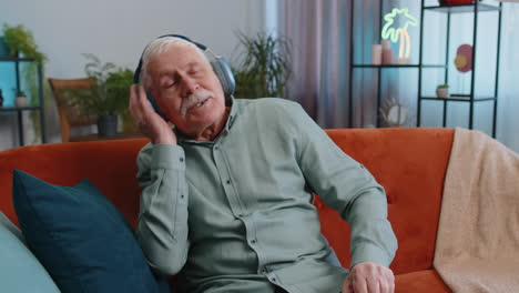 Feliz-Abuelo-Con-Auriculares-Inalámbricos-Relajándose-Se-Sienta-En-El-Sofá-De-Casa-Escuchando-Su-Música-Favorita