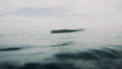 Small atoll tropical island off in distance behind calm ocean water surface