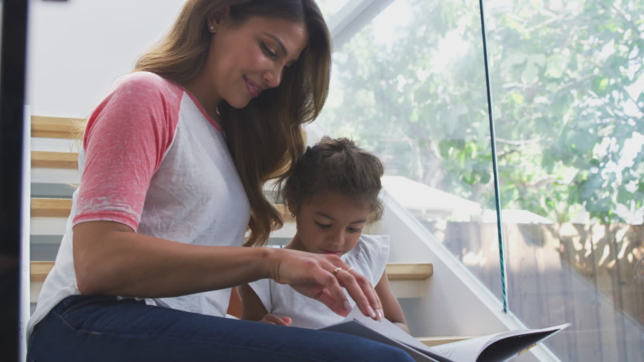 madre y hija hispanas sentadas en la escalera en una casa moderna leyendo un libro juntas