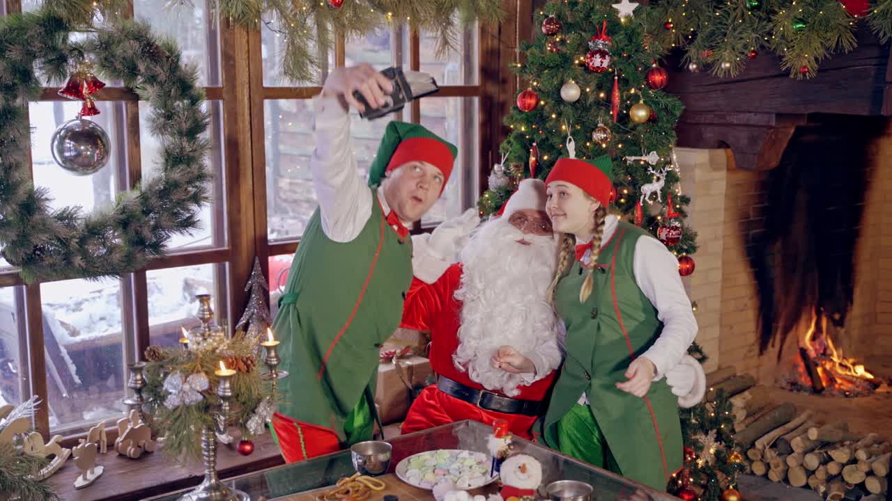 Happy elves and Santa near Christmas tree. Santa Claus together with joyful elves making photo. Fairy characters doing selfie at Christmas.