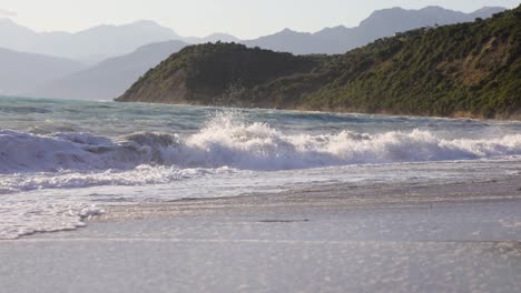 Waves-crushing-on-a-beach-in-slow-motion-with-cliffside-and-mountains-on-the-background