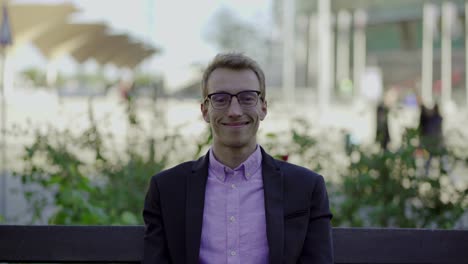 Smiling-young-man-in-eyeglasses-sitiing-on-wooden-bench-in-park