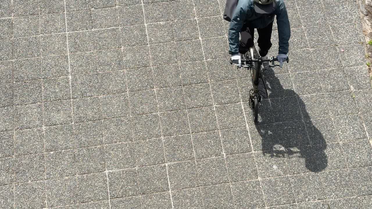 A cyclist wearing a helmet and backpack rides across stone pavement in bright morning light