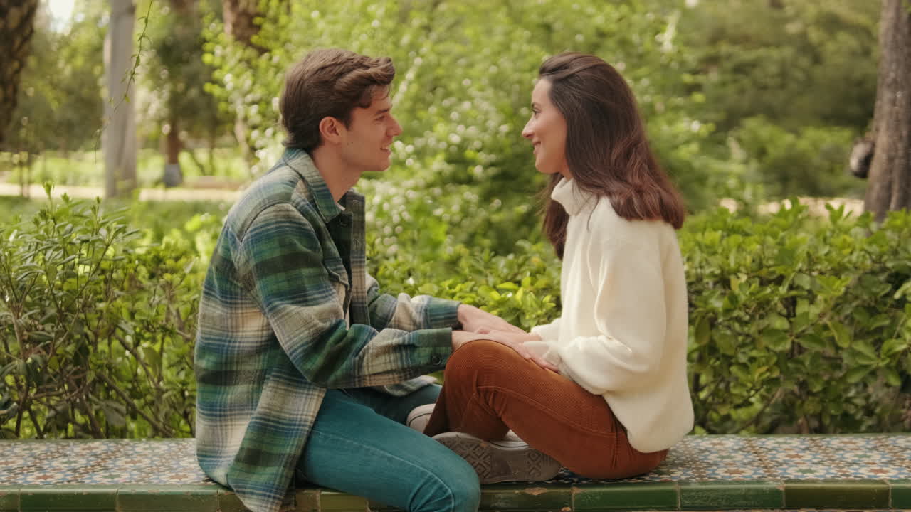 Happy young couple man and woman laughing kissing holding hands sitting in park