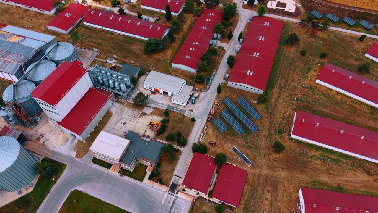 Landbrugsforarbejdningsanlæg med siloer og solpaneler. Skråt luftfoto af røde tagkonstruktioner, lagertanke, maskineri, veje og tørt græs omkring anlægget.