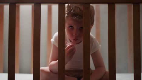 Blond-toddler-sits-in-wooden-crib-eating-slice-of-sausage