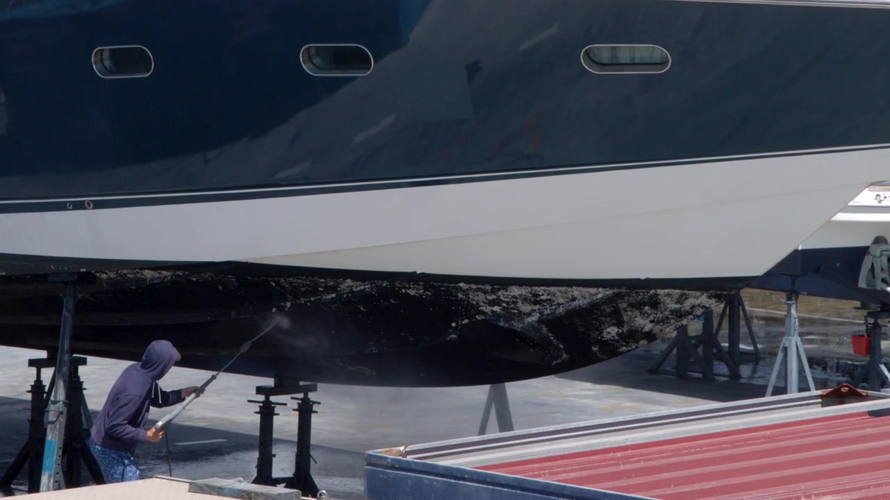 Caucasian worker uses high-pressure washer on superyacht hull during maintenance in sunny French shipyard.