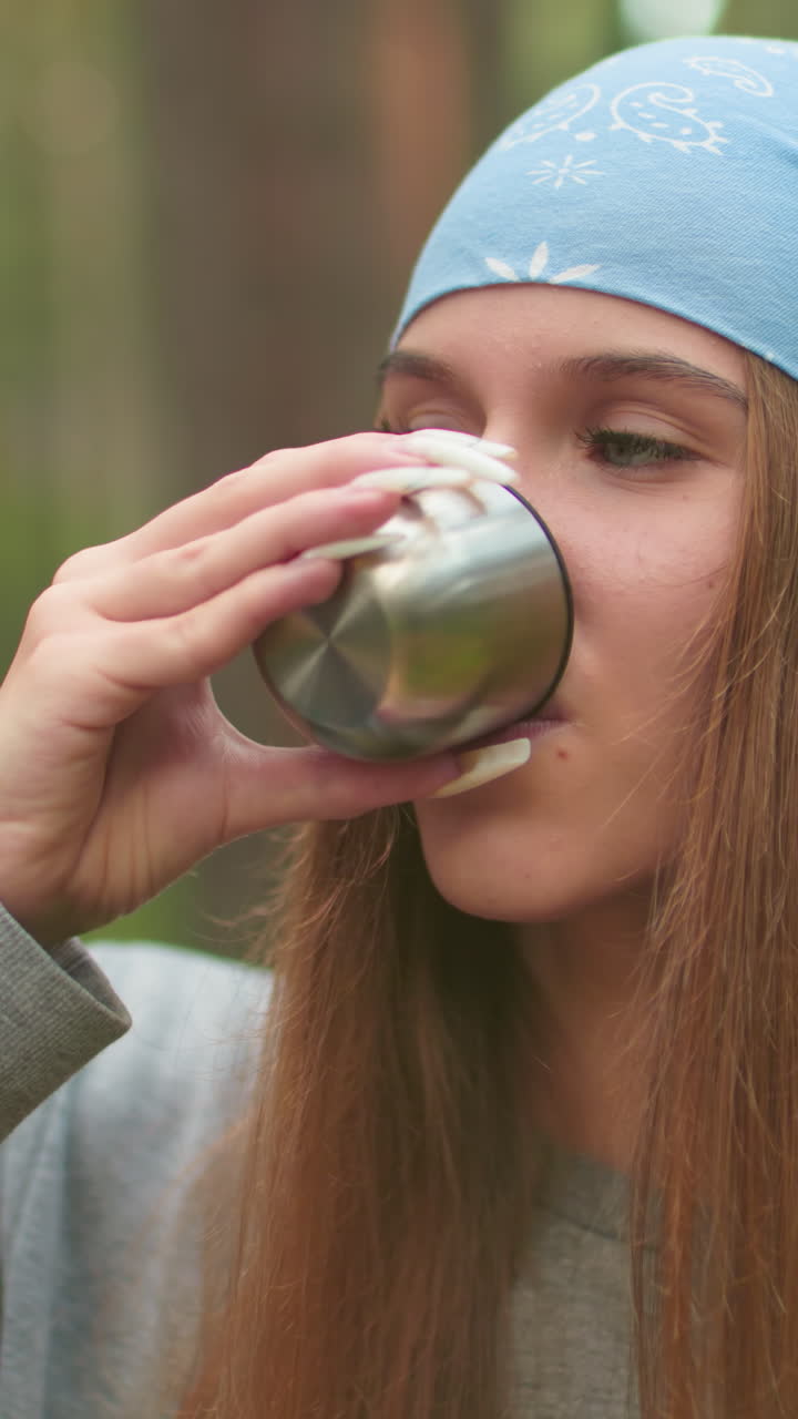 primer plano de una mujer joven con pañuelo azul bebiendo agua de una taza térmica en un bosque verde sereno, centrándose en su expresión tranquila y relajada, con uñas delicadas y bien cuidadas